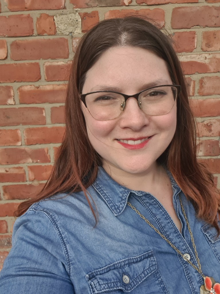 Melissa Wilson Craw, therapist for children and young people, smiles. She has dark hair, light skin and brown eyes, and she stands in from of a brick wall wearing a denim top and a gold chain with a coral coloured flower on it